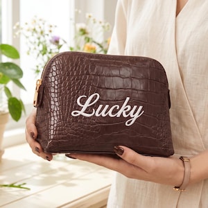 May include: A dark brown cosmetic bag with a crocodile-embossed texture and a gold-toned zip. The word Lucky is written in white cursive script across the centre of the front panel. The bag has a rounded top and a structured, semi-circular shape.
