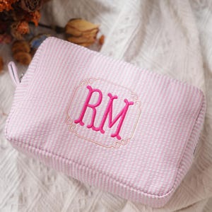 May include: A pink and white striped cosmetic bag with a rectangular embroidered frame. The frame contains the pink embroidered letters "RM". The bag has a zip closure and a small loop on the side.