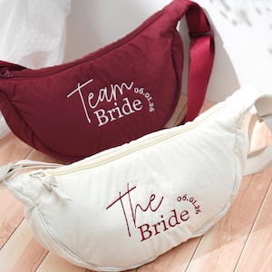 Może przedstawiać: Dwie nerki, jedna bordowa i jedna biała, z haftowanym tekstem. Bordowa nerka ma napis "Team Bride 06.01.26" białym pismem. Biała nerka ma napis "The Bride 06.01.26" bordowym pismem. Obie mają zamki błyskawiczne.