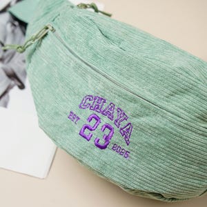 May include: A mint green corduroy fanny pack with a zipper closure and drawstring. The bag features the embroidered text "CHAYA 23 2025" in purple. The bag is on a neutral background.
