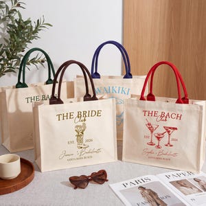 May include: Four off-white canvas tote bags with colored handles and printed designs. One features a gold cowboy boot and text reading THE BRIDE CLUB EST 2026. Another has red cocktail glasses and THE BACH CLUB text. Others show green and blue text.