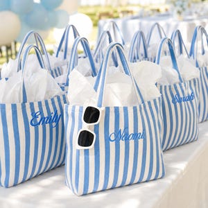 May include: A collection of blue and white striped canvas tote bags with two handles, each filled with white tissue paper. The bags feature embroidered names such as Emily, Naomi, and Sarah in blue script. A pair of white sunglasses hangs on one bag.