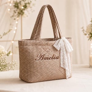 Könnte beinhalten: Eine gesteppte Tragetasche mit braun-weißem Vichy-Muster. Auf der Vorderseite ist der Name Amelia in brauner Schreibschrift eingestickt. Ein weißes Spitzenband ist an der rechten Seite der Tasche befestigt, die zwei passende Stoffgriffe hat.