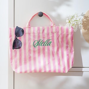 Puede incluir: Un bolso tote a rayas rosas y blancas con el nombre "Stella" en escritura verde. Gafas de sol negras cuelgan del lateral de la bolsa. La bolsa tiene una capa de pl&aacute;stico transparente.