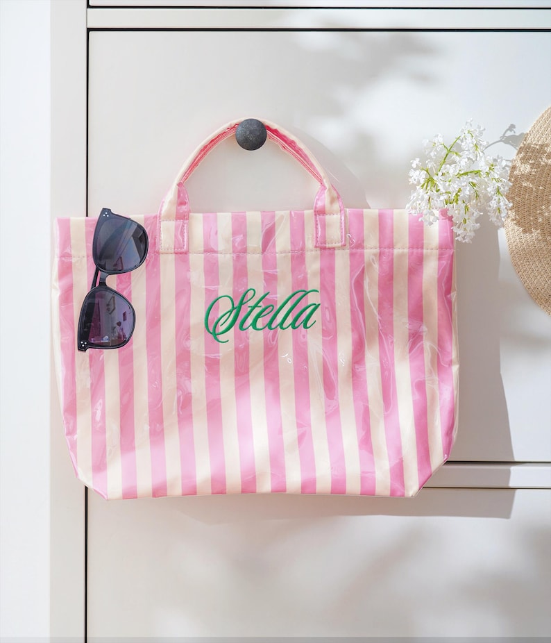 Puede incluir: Un bolso tote a rayas rosas y blancas con el nombre "Stella" en escritura verde. Gafas de sol negras cuelgan del lateral de la bolsa. La bolsa tiene una capa de pl&aacute;stico transparente.