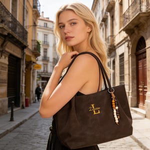 Könnte beinhalten: Eine braune Tragetasche mit dem Namen "Louise" in Gold gestickt. Die Tasche hat eine dekorative Quaste mit Holzperlen und einem Metallring. Die Tasche wird über der Schulter getragen.