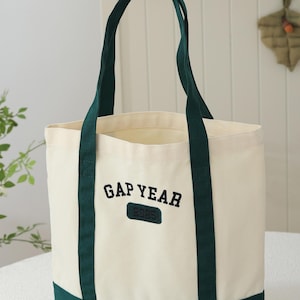 May include: Cream-colored tote bag with dark green accents. The bag features dark green handles and a dark green base. The text "GAP YEAR" is embroidered in black on the front, with "2025" below it.