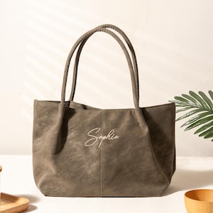 Könnte beinhalten: Eine taupefarbene Tragetasche mit zwei Henkeln. Die Tasche trägt den Namen "Sophia" in kursiver Schrift gestickt. Die Tasche ist aus einem weichen, strukturierten Material gefertigt und hat ein schlichtes, elegantes Design, das für den täglichen Gebrauch geeignet ist.