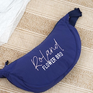 Op de afbeelding: Een marineblauwe heuptas met de naam "Roland" en de woorden "FLOWER BRO" in witte letters. De tas heeft een halvemaanvorm en een marineblauwe riem. De achtergrond is een beige geweven mat.