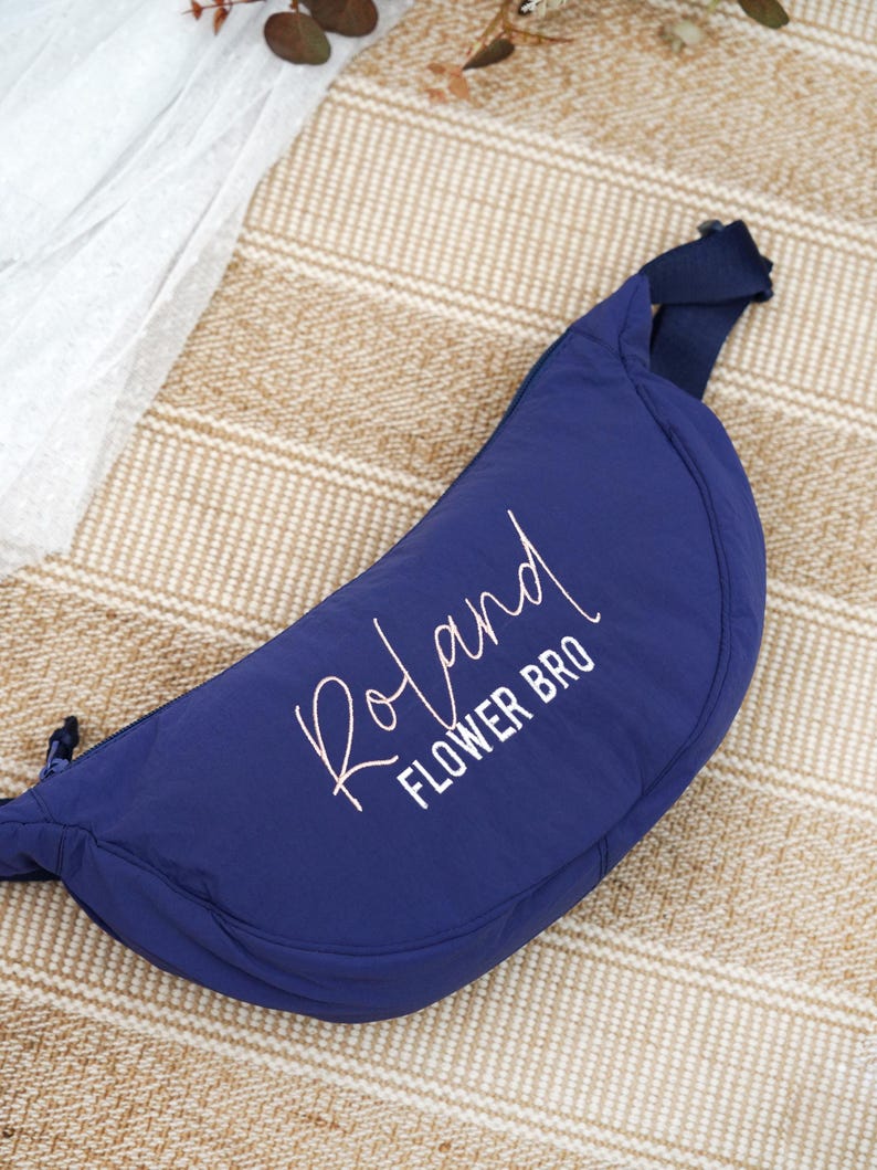 Op de afbeelding: Een marineblauwe heuptas met de naam "Roland" en de woorden "FLOWER BRO" in witte letters. De tas heeft een halvemaanvorm en een marineblauwe riem. De achtergrond is een beige geweven mat.