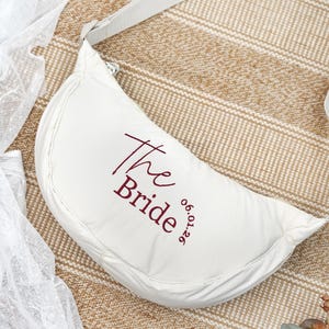 Op de afbeelding: Een witte, halvemaanvormige tas met een bijpassende riem. De tekst "The Bride" is geborduurd in bordeauxrood, samen met de datum "06.01.26". De tas is waarschijnlijk voor een bruiloft of bruidsevenement.