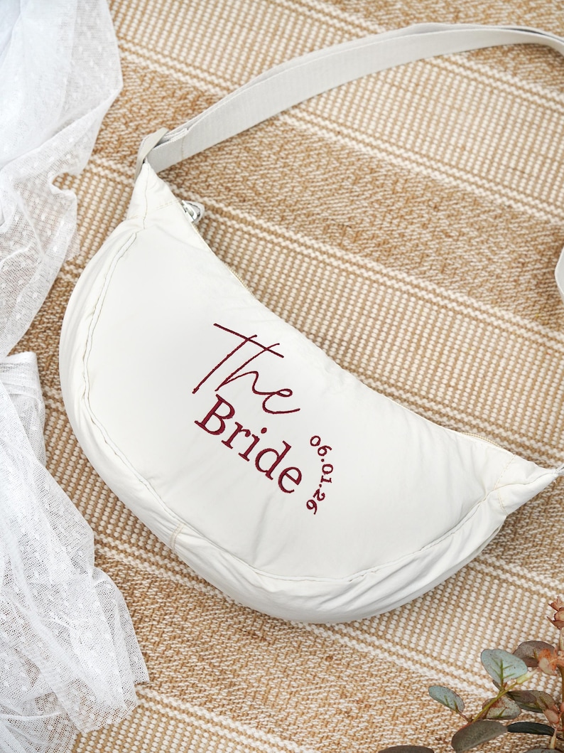 Op de afbeelding: Een witte, halvemaanvormige tas met een bijpassende riem. De tekst "The Bride" is geborduurd in bordeauxrood, samen met de datum "06.01.26". De tas is waarschijnlijk voor een bruiloft of bruidsevenement.
