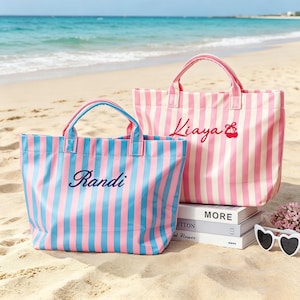 Puede incluir: Dos bolsos tote a rayas en una playa de arena. Un bolso es azul y rosa con el nombre "Randi" bordado. El otro bolso es rosa y blanco con el nombre "Kiaya" y un dise&ntilde;o de cereza. Los bolsos tienen asas a juego.
