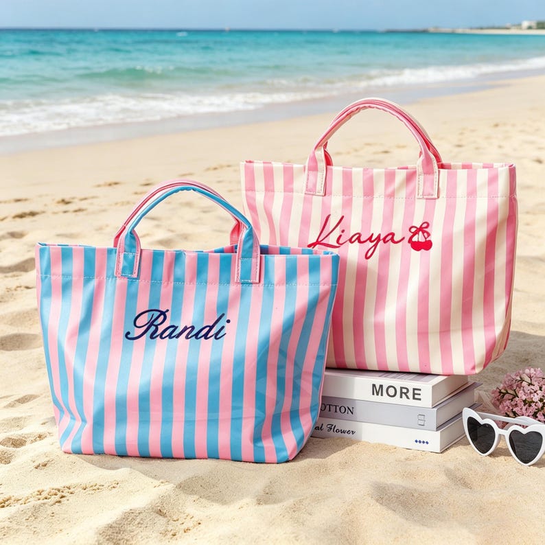 Puede incluir: Dos bolsos tote a rayas en una playa de arena. Un bolso es azul y rosa con el nombre "Randi" bordado. El otro bolso es rosa y blanco con el nombre "Kiaya" y un dise&ntilde;o de cereza. Los bolsos tienen asas a juego.
