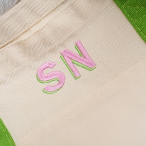 May include: Close-up of a canvas tote bag with the embroidered letters "SN". The letters are pink with a green outline. The bag has a cream-coloured body and green trim.