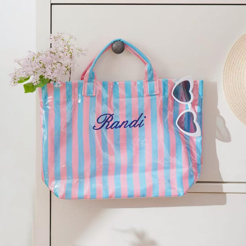 Puede incluir: Un bolso tote transparente con rayas verticales rosas y azules. El bolso tiene un asa rosa y el nombre "Randi" en escritura cursiva. Gafas de sol blancas en forma de coraz&oacute;n est&aacute;n unidas al bolso.