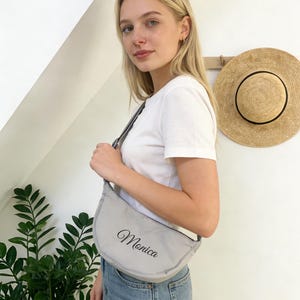 Op de afbeelding: Een lichtgrijze, halvemaanvormige schoudertas met de naam "Monica" in elegant schrift. De tas wordt over de schouder gedragen. Een strohoed hangt op de achtergrond aan de muur.