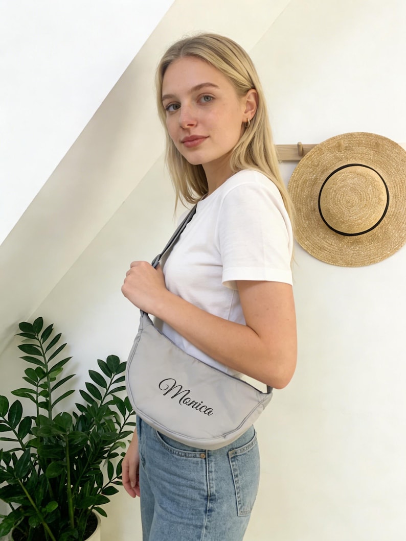 Op de afbeelding: Een lichtgrijze, halvemaanvormige schoudertas met de naam "Monica" in elegant schrift. De tas wordt over de schouder gedragen. Een strohoed hangt op de achtergrond aan de muur.