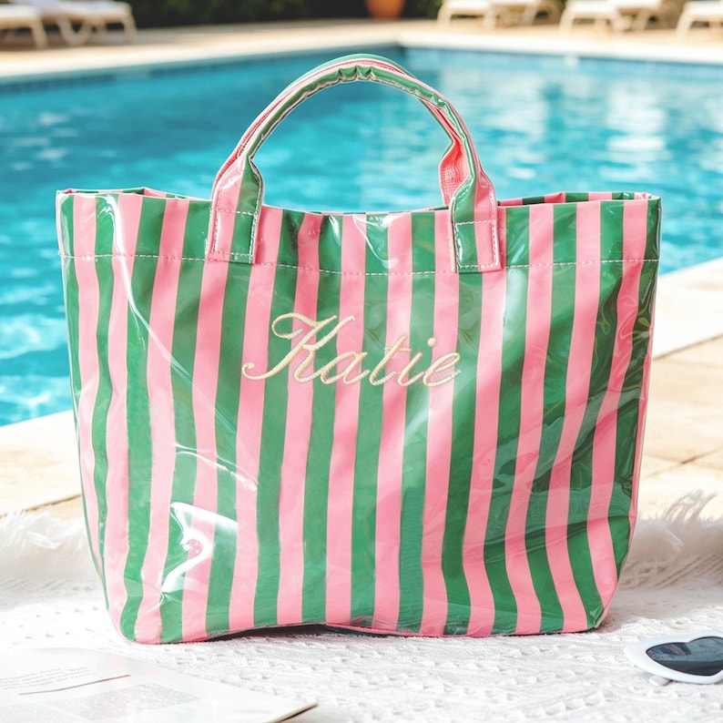 Puede incluir: Un bolso tote a rayas rosas y verdes con el nombre "Katie" bordado en cursiva. El bolso tiene un acabado transparente y brillante y un asa a juego. El bolso est&aacute; cerca de una piscina.
