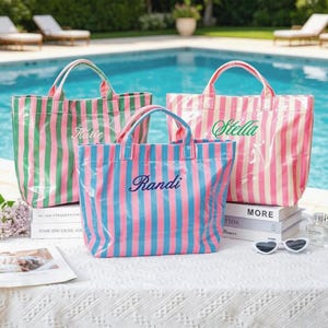 Puede incluir: Tres bolsos tote a rayas en verde y rosa, azul y rosa, y rosa y blanco. Cada bolso tiene un nombre bordado: Katie, Randi y Stella. Los bolsos tienen asas y est&aacute;n sobre una superficie blanca.