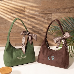 Könnte beinhalten: Zwei Wildleder-Schultertaschen in Grün und Braun. Jede Tasche hat einen passenden Henkel und einen dekorativen Schal, der am Henkel befestigt ist. Die grüne Tasche hat den Buchstaben "T" aufgestickt, und die braune Tasche hat die Buchstaben "L/R".