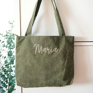 Könnte beinhalten: Olivgrüne Cord-Einkaufstasche mit langen Trägern. Das Wort "Maria" ist in Schreibschrift auf die Vorderseite der Tasche gestickt. Die Tasche hat einen Reißverschluss und hängt vor einem weißen Hintergrund.