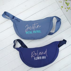Op de afbeelding: Twee blauwe heuptassen met verstelbare bandjes. De bovenste tas is lichtblauw met de naam "Justin" en de woorden "Petal Patrol" in een scriptlettertype. De onderste tas is marineblauw met de naam "Roland" en de woorden "Flower Bro".