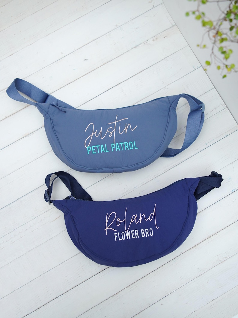 Op de afbeelding: Twee blauwe heuptassen met verstelbare bandjes. De bovenste tas is lichtblauw met de naam "Justin" en de woorden "Petal Patrol" in een scriptlettertype. De onderste tas is marineblauw met de naam "Roland" en de woorden "Flower Bro".