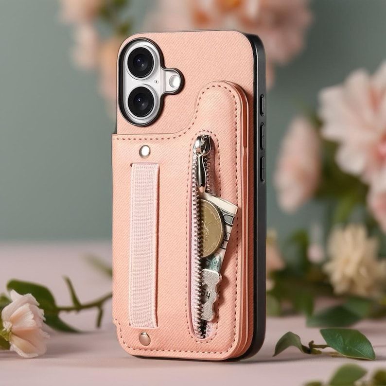 May include: A pink phone case with a wallet feature. The case has a zipper pocket containing a key, a coin, and a bill. A pink strap is attached to the case. The phone case is shown against a blurred floral background.