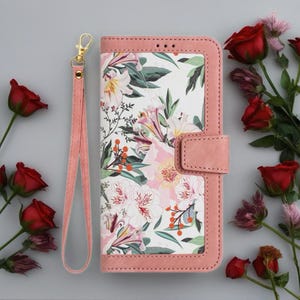 May include: A pink floral phone case with a matching wrist strap. The case has a white background with pink and green floral patterns. The edges and strap are a soft pink colour, and the strap has a gold-coloured clasp.