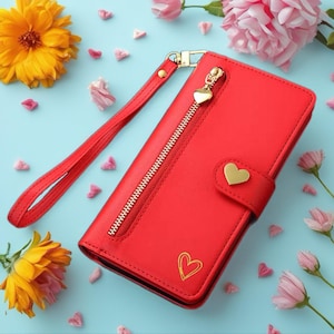 May include: A red wallet with a gold zip and heart-shaped zip pull. The wallet has a wrist strap and a gold heart-shaped clasp. A gold heart outline is on the front. The wallet is surrounded by flowers and heart-shaped confetti.