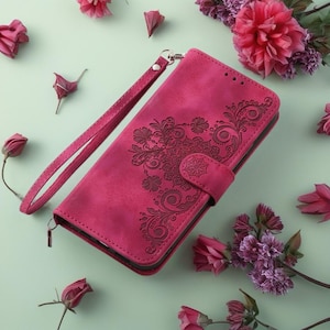 May include: A magenta phone wallet with an embossed floral design. The wallet features a wrist strap and a magnetic clasp. The design includes a central mandala-like pattern with swirling floral elements. Pink flowers surround the wallet.