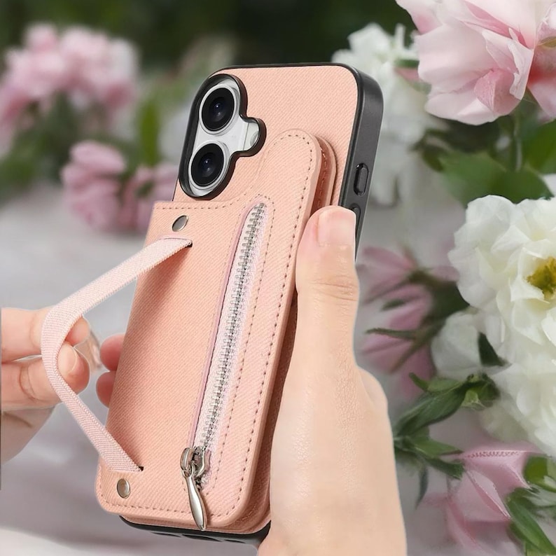 May include: A pink phone case with a black border and a matching wallet attachment. The wallet has a zipper closure and a wrist strap. The phone case is shown against a background of pink and white flowers.