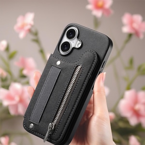 May include: Black phone case with a wallet attachment. The case has a zippered pocket and a fabric hand strap. The case is held in front of a blurred background of pink flowers.