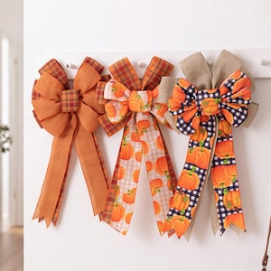 May include: Three decorative bows made of fabric ribbon. The left bow is solid orange with plaid edges. The middle bow features a light orange gingham pattern with pumpkin prints. The right bow has a black and white buffalo check with pumpkins.
