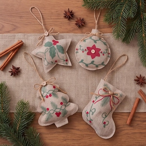 Puede incluir: Cuatro adornos colgantes de tela color canela, que incluyen una estrella, un círculo, un árbol y un calcetín. Cada pieza presenta follaje verde bordado, bayas rojas y un pequeño botón de madera. Los adornos están suspendidos por cordeles.