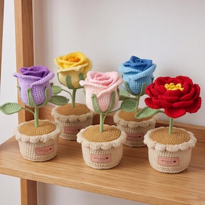 May include: Five crocheted flowers in pots, each featuring a unique color: purple, yellow, pink, blue, and red. Each flower has green stems and leaves, and is planted in a beige crocheted pot with a scalloped rim and a small rectangular label.