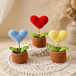 May include: Three crochet heart-shaped flowers in brown pots. One is red, one is blue, and one is yellow. Each flower has green leaves and a green stem. The pots are sitting on a white, textured surface.