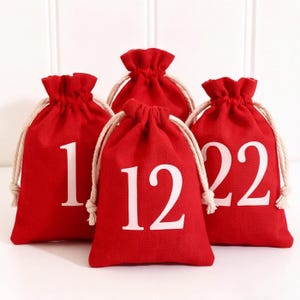 Puede incluir: Cuatro bolsas de tela de color rojo brillante con cordones blancos. Cada bolsa presenta un número grande impreso en blanco en la parte frontal. Los números visibles son 1, 12 y 22, con una bolsa parcialmente oculta detrás.