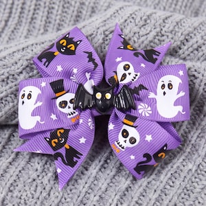 Könnte beinhalten: Eine violette Haarschleife aus Ripsband mit einem wiederkehrenden Muster aus weißen Geistern, schwarzen Katzen und Totenköpfen mit Zylinderhüten, in der Mitte verziert mit einer schwarzen Fledermaus aus Kunststoff mit gelben Augen.