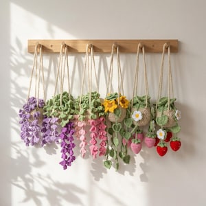 May include: Six crocheted hanging planters suspended from a light wood wall rack. The planters feature various designs including purple wisteria, pink flowers, yellow sunflowers, green vines, and red strawberries with white flowers, all crafted from yarn.