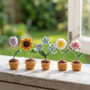 Puede incluir: Cinco flores de ganchillo en macetas pequeñas. Las flores incluyen un tulipán morado, un girasol amarillo, nomeolvides azules, una margarita blanca y una rosa rosa. Las macetas son amarillas con tierra marrón, y los tallos y hojas son verdes.