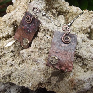 May include: A pair of copper earrings with a hammered texture and a swirl design. The earrings are attached to silver hooks.