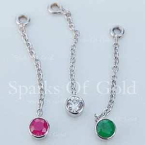 May include: Three silver chain earrings, each with a different colored gemstone: a ruby, a clear diamond, and an emerald. The earrings are displayed on a light background, with the text "Sparks Of Gold" visible.