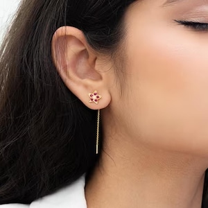 May include: A close-up of a person wearing a gold earring with a small, red flower design. The earring has a long, thin gold chain that dangles down. The person has dark hair and is wearing a white top.