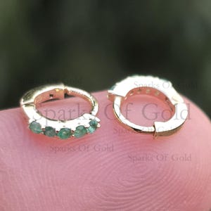 Puede incluir: Pendientes de aro dorados con pequeñas piedras preciosas redondas de color verde esmeralda. Un pendiente tiene cinco esmeraldas, el otro tiene piedras más pequeñas. Los pendientes se muestran sobre una superficie clara.