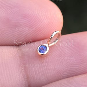 Op de afbeelding: Een kleine, gouden hanger met een ronde, blauwe edelsteen. De hanger heeft een eenvoudig ontwerp met een lus voor een ketting. De edelsteen is gezet in een gouden bezel.