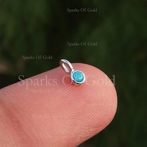 May include: A small, silver pendant with a round, turquoise-coloured stone. The pendant has a loop at the top for a chain. The background is a blurred green.