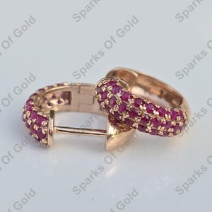 Pode incluir: Brincos de argola em ouro rosa cravejados com pequenas e vibrantes pedras preciosas vermelhas. Os brincos têm um fecho seguro e um acabamento polido. A joia é apresentada sobre um fundo macio e neutro.