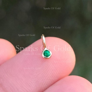May include: A small, gold pendant featuring a round, vivid green gemstone. The pendant is held between fingers, with the text "Sparks Of Gold" visible in the background. The gemstone is the central feature.